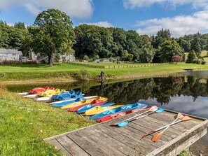 A row of numbered kayaks and paddles docked at a wooden pier by a calm lake, surrounded by lush greenery and a wooden fence. - 4 Bed in Bala (oc-75288) (Llandderfel)