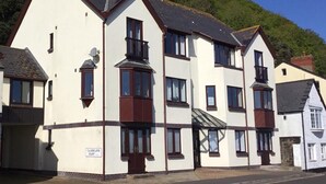 A row of white buildings with dark brown trim, a sign indicating a court, and a hillside in the background. - 2 Bed in Minehead (oc-75338) (Minehead)