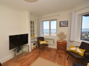 A living room with a TV, two chairs, a wooden side table, and a window with a view of the sea.