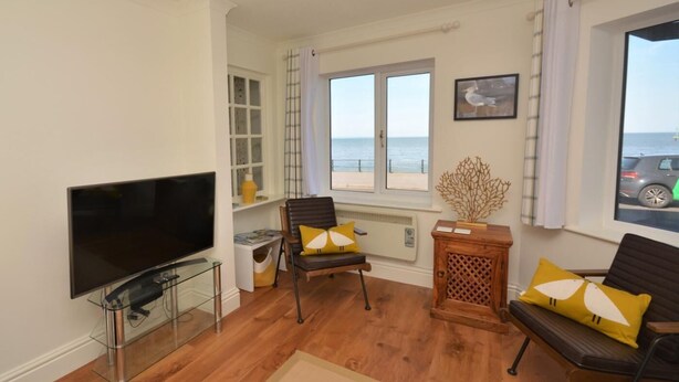 A living room with a TV, two chairs, a wooden side table, and a window with a view of the sea. - 2 Bed in Minehead (oc-75338) (Minehead)
