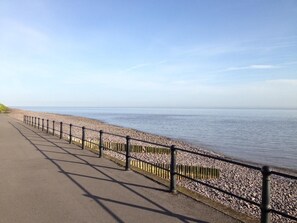 A paved walkway alongside a pebble beach with a metal railing.