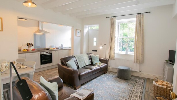 Ett modernt vardagsrum med en brun lädersoffa, ett matbord med stolar, en köksdel och ett stort fönster med gardiner.