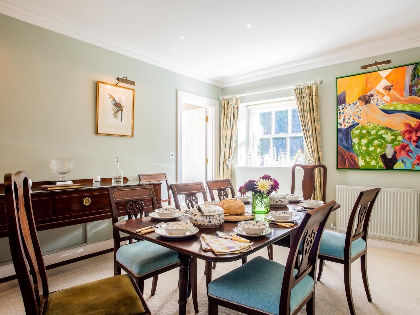 Une salle à manger avec une table mise pour quatre personnes, des chaises, un vase rempli de fleurs et un tableau accroché au mur.