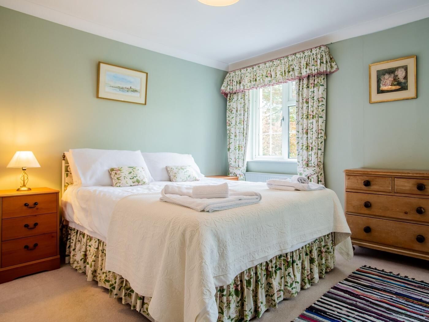 Una cama bien hecha con una colcha floral, una lámpara de noche, un aparador de madera y una ventana con cortinas estampadas.