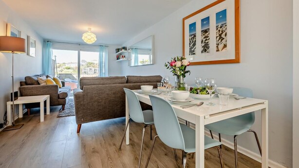 A modern living room with a dining area, a sofa set, a table with chairs, and a large window. - 2 Bed in Newquay (oc-75578) (Newquay)