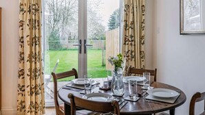 Ein Essbereich mit einem runden Tisch, gedeckt für vier Personen, einer Blumenvase und Blick auf einen Garten durch Glastüren.