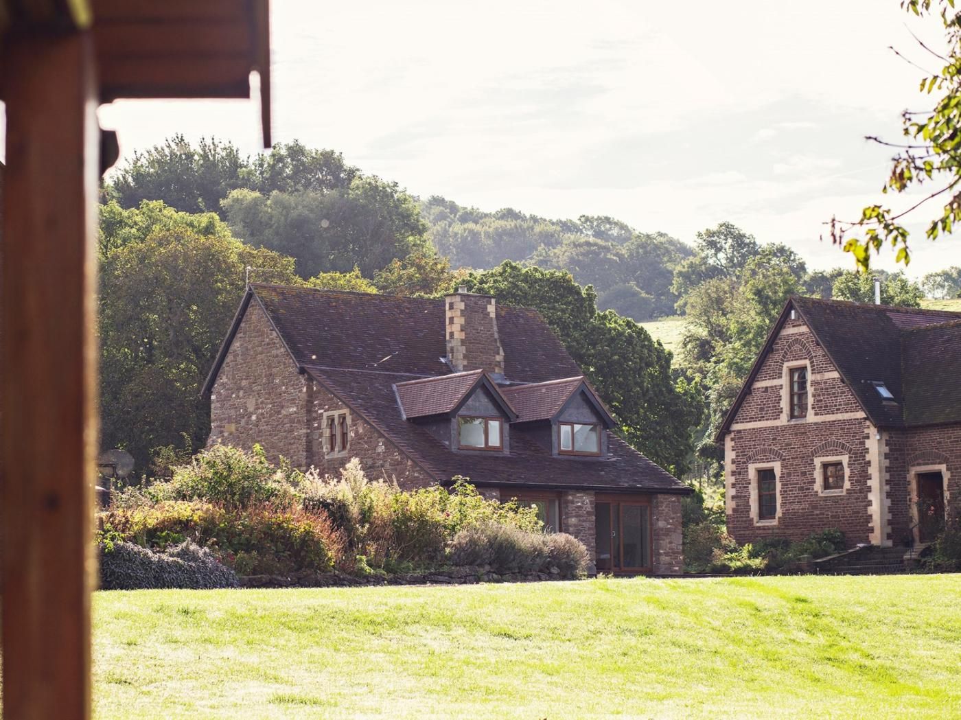 Une maison traditionnelle en pierre, dotée d’un toit en pente, entourée d’un champ verdoyant et d’une forêt dense.