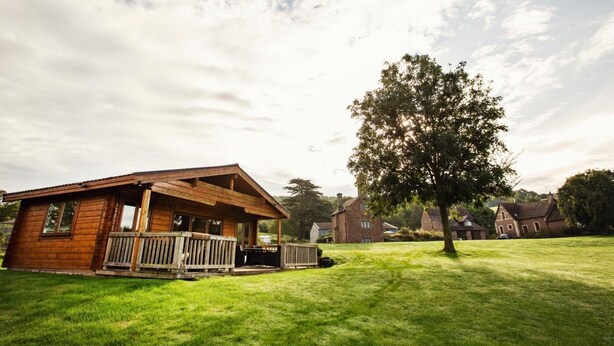 A wooden cabin with a porch, surrounded by a grassy lawn and a large tree. - 4 Bed in Worcester (oc-75601) (Halmond's Frome)
