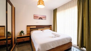 Hotellihuone, jossa on sänky, yöpöytä, peili, taulu, ikkuna verhoilla ja kattovalaisin.