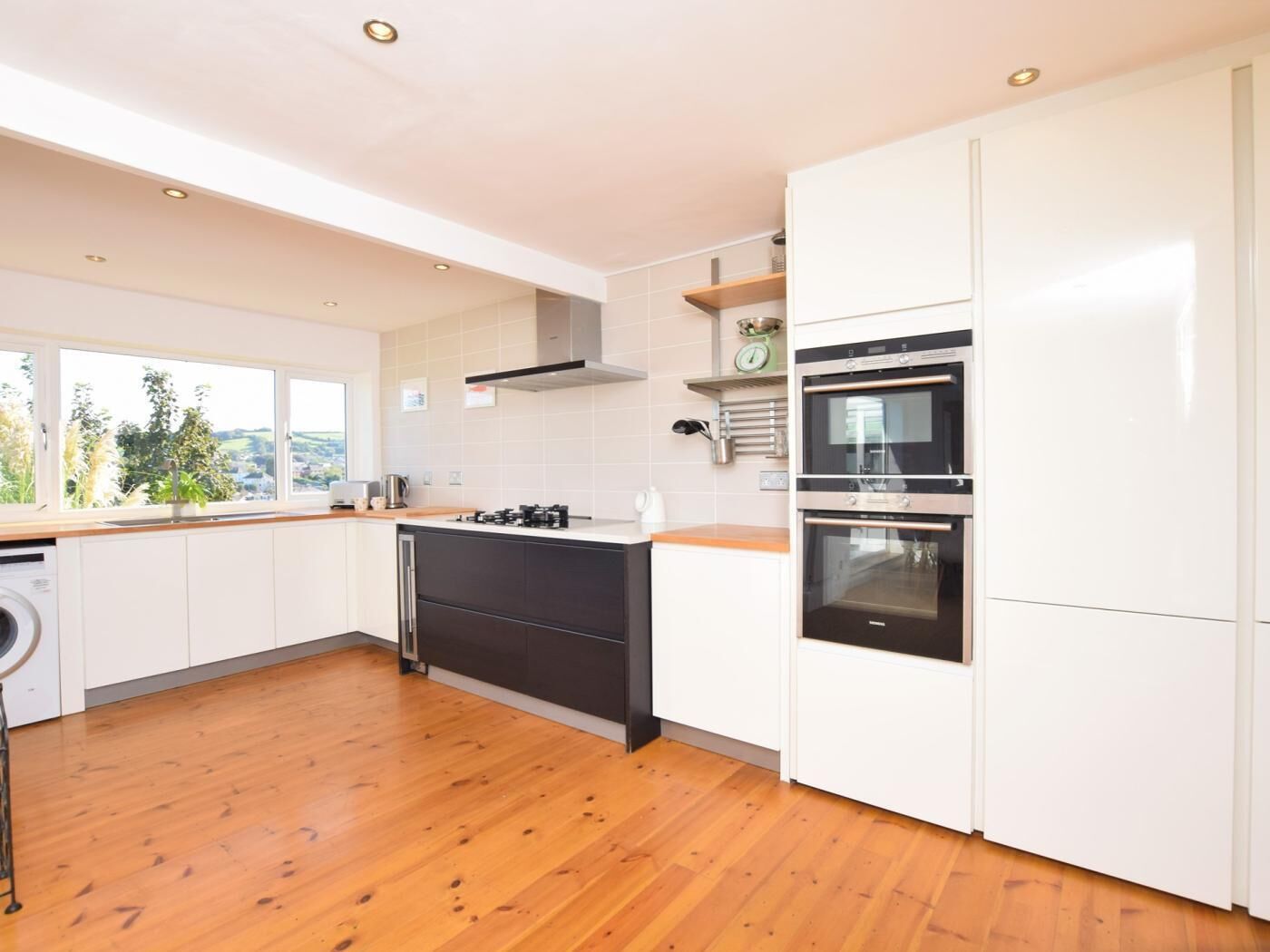 A modern kitchen with white cabinets, a wooden floor, a built-in oven, and a window overlooking greenery.