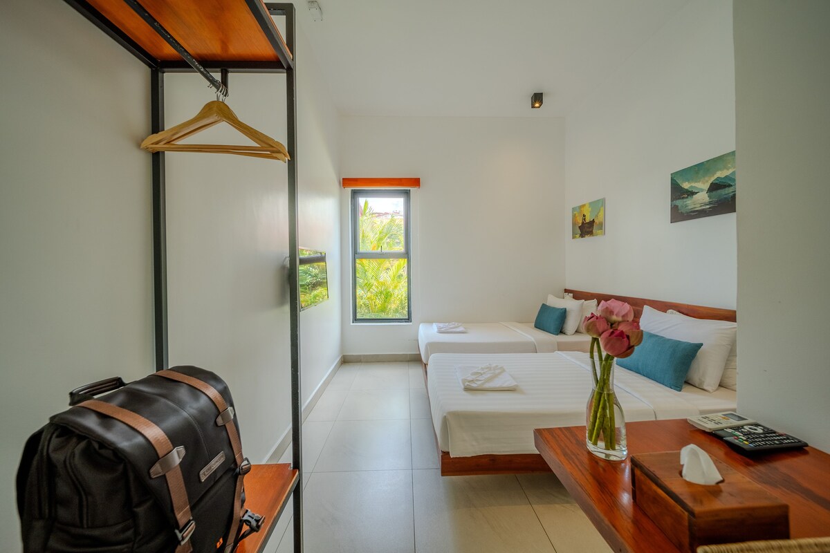 A hotel room with two beds, a wooden desk, a hanging coat rack, and a window with a view of greenery.