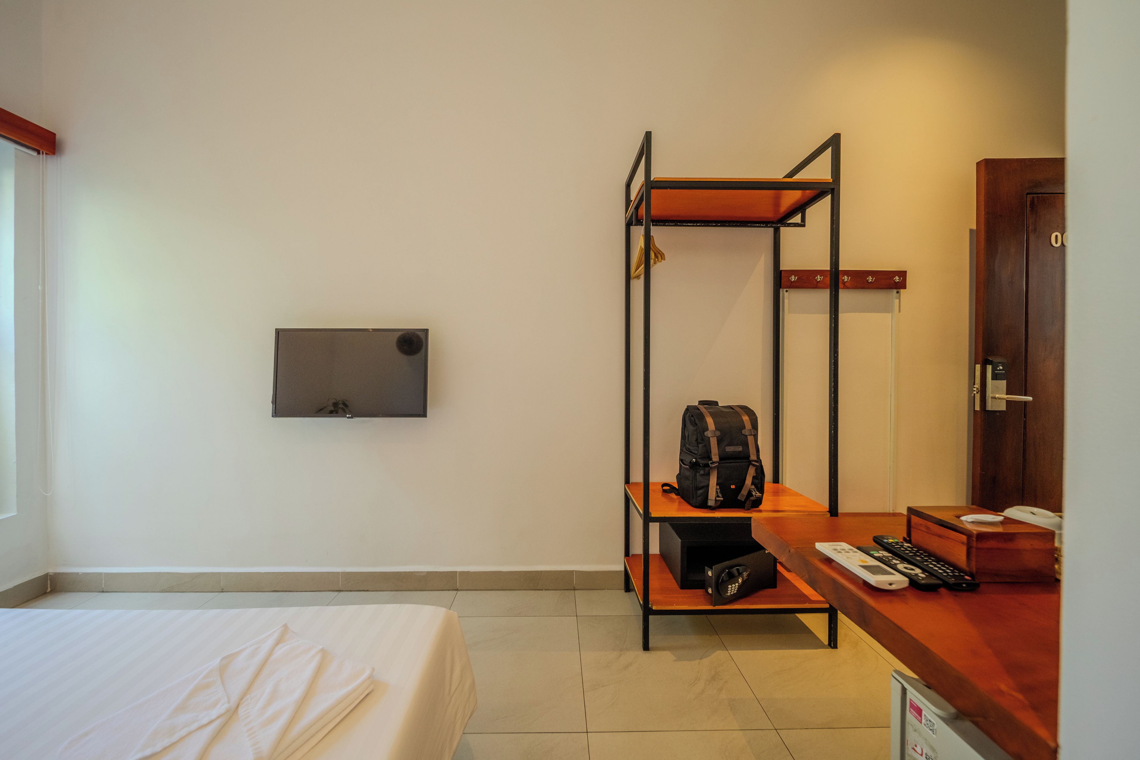 Habitación de hotel con cama, escritorio con silla, estante con una bolsa, perchero y una puerta.