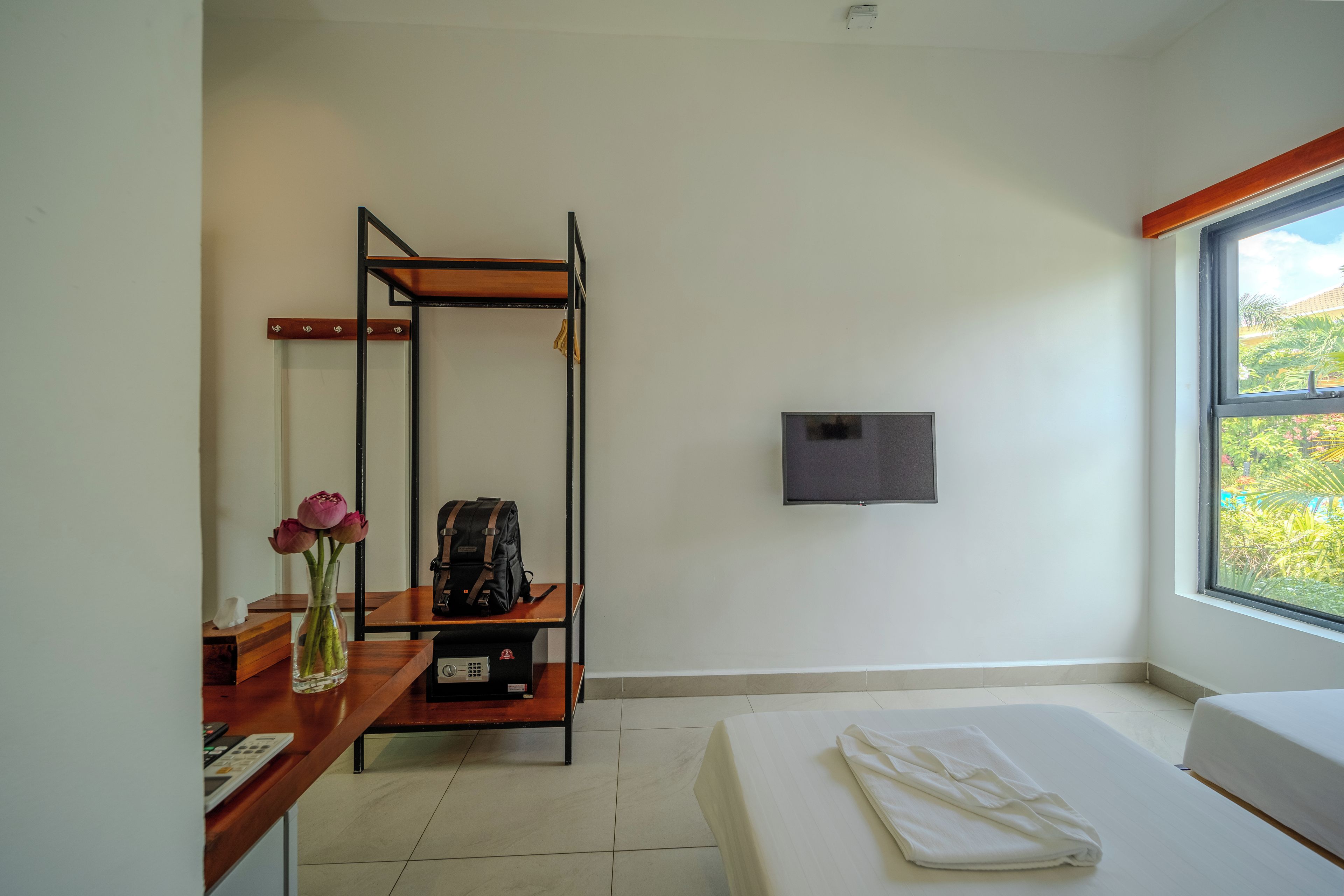 Habitación de hotel con cama, escritorio con laptop, pequeña mesa con un florero y televisor montado en la pared, además de ventana con vista a follaje.