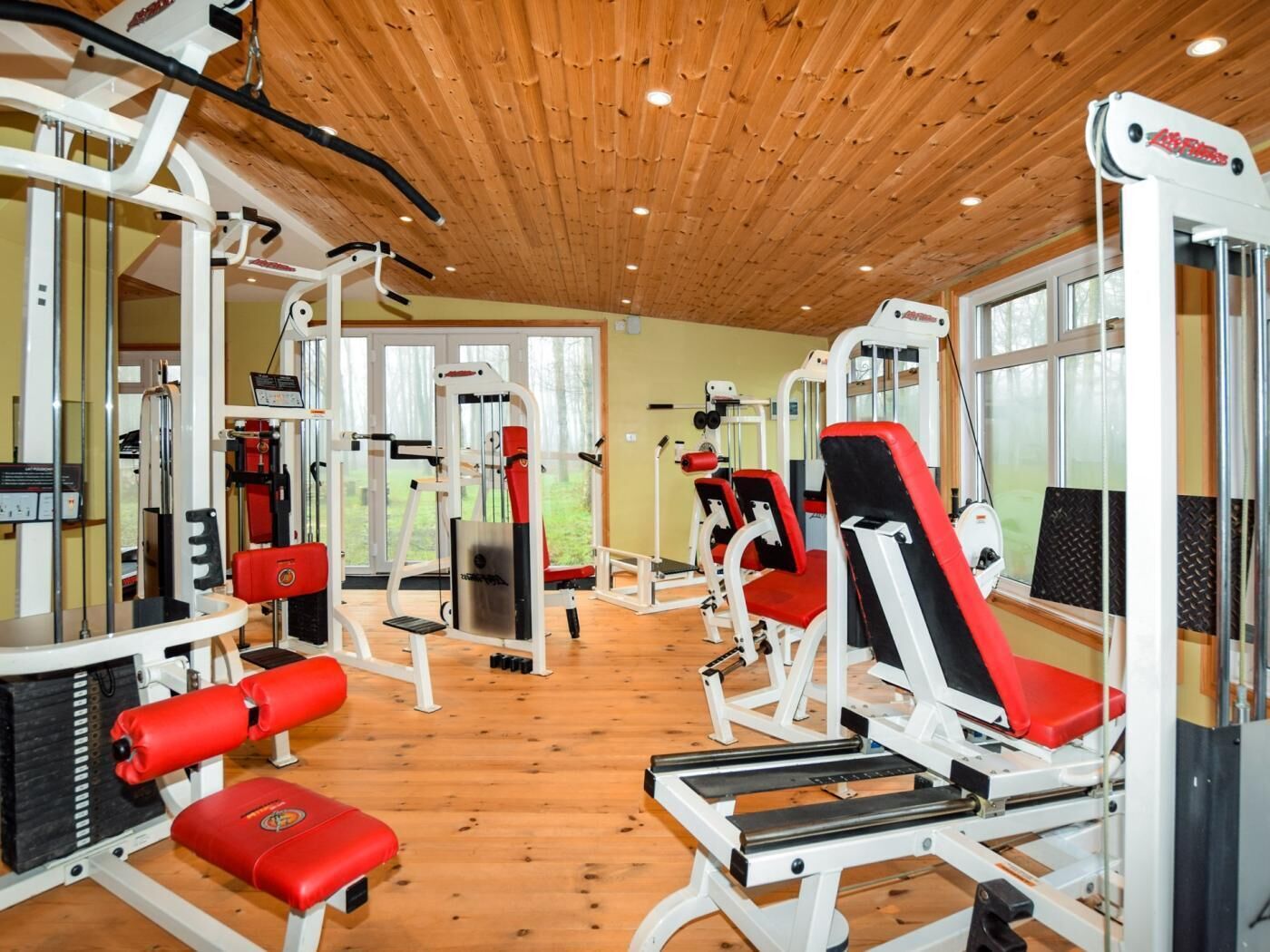 A gym with various exercise machines, wooden flooring, and large windows.