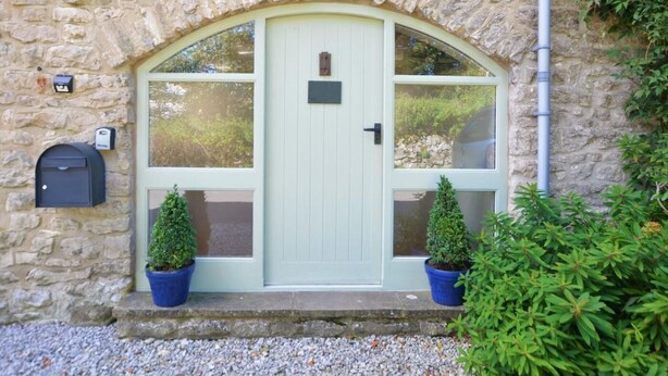 Un bâtiment en pierre avec une porte verte, deux plantes en pot, une boîte aux lettres et une caméra de surveillance.