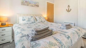 Een slaapkamer met een bed, nachtkastje, lamp, schilderij, deur en een anker decoratie.