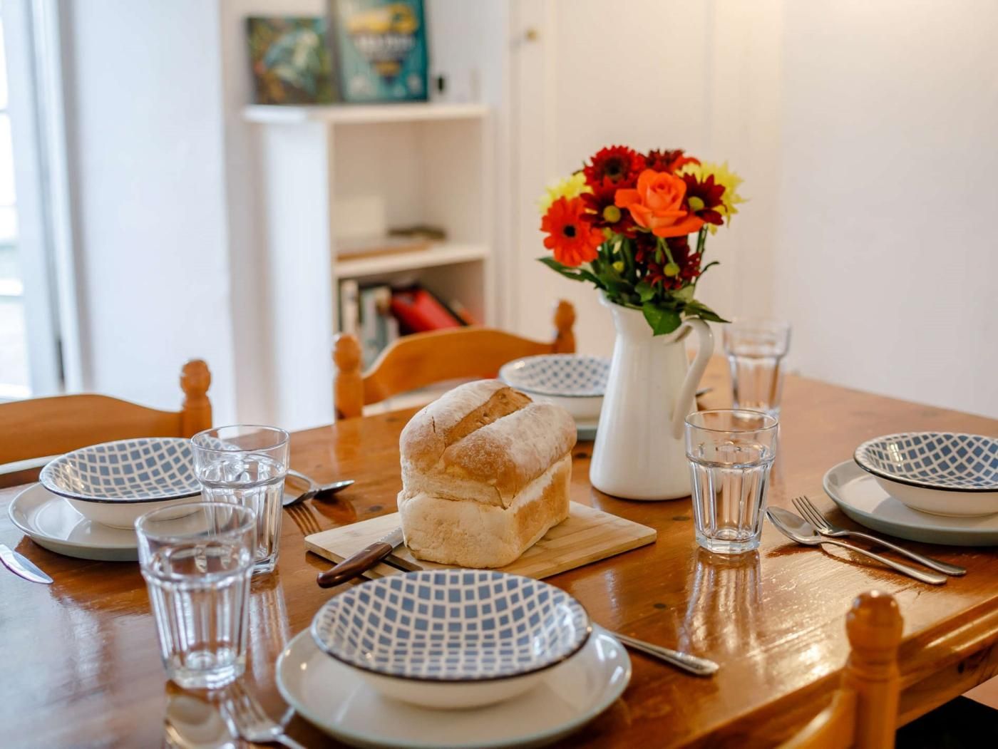 A dining table set with plates, glasses, cutlery, a loaf of bread, and a vase with flowers.
