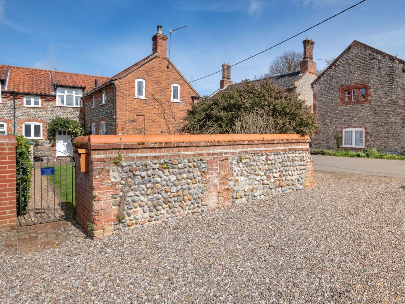 A traditional stone wall with a gravel driveway, a metal gate, and a brick building with a tiled roof.