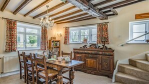 Une salle à manger traditionnelle avec une table en bois, des chaises et un lustre. Un escalier se trouve à droite, et il y a une fenêtre avec des rideaux.