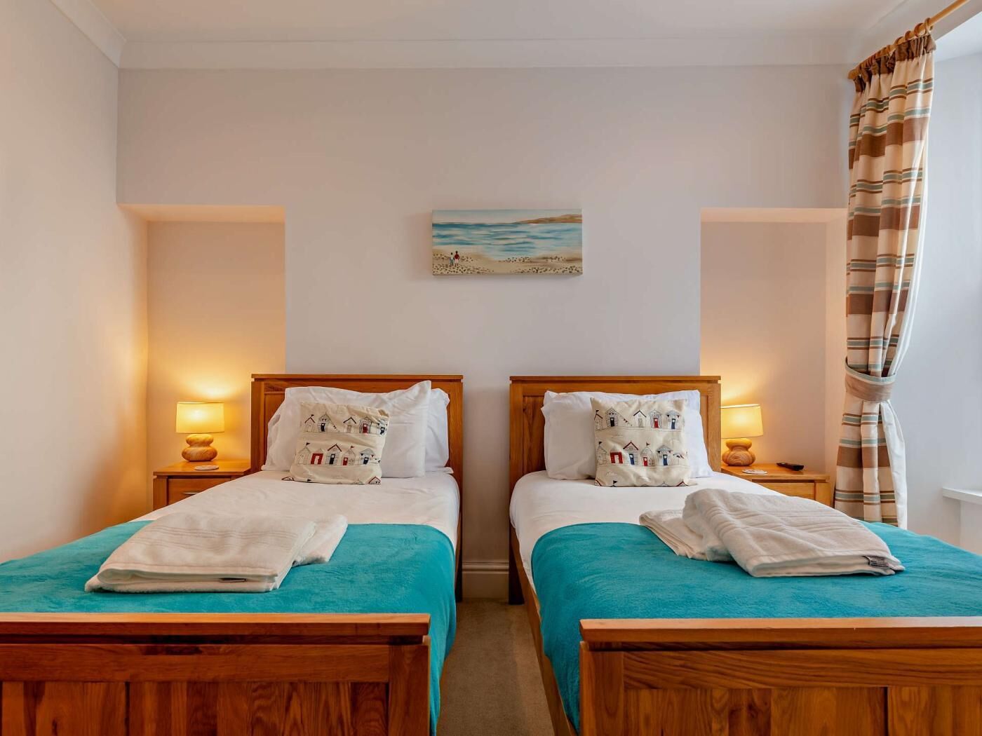 A double room with two beds, each with a wooden headboard, a teal bedspread, and matching pillows. There are bedside lamps, a painting on the wall, and a window with curtains.