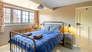 Een slaapkamer met een metalen bedframe, een blauw gebogen beddeken, twee bedlampen, een radiator, een raam met gordijnen, en een deur.
