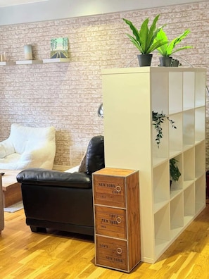 Een woonkamer met een bakstenen muur, een bank, een zwarte leren fauteuil, een houten kast met lades en een witte boekenkast met planten.