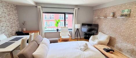 A hotel room with a large bed, a desk, a chair, a TV, and a window with a view of buildings.