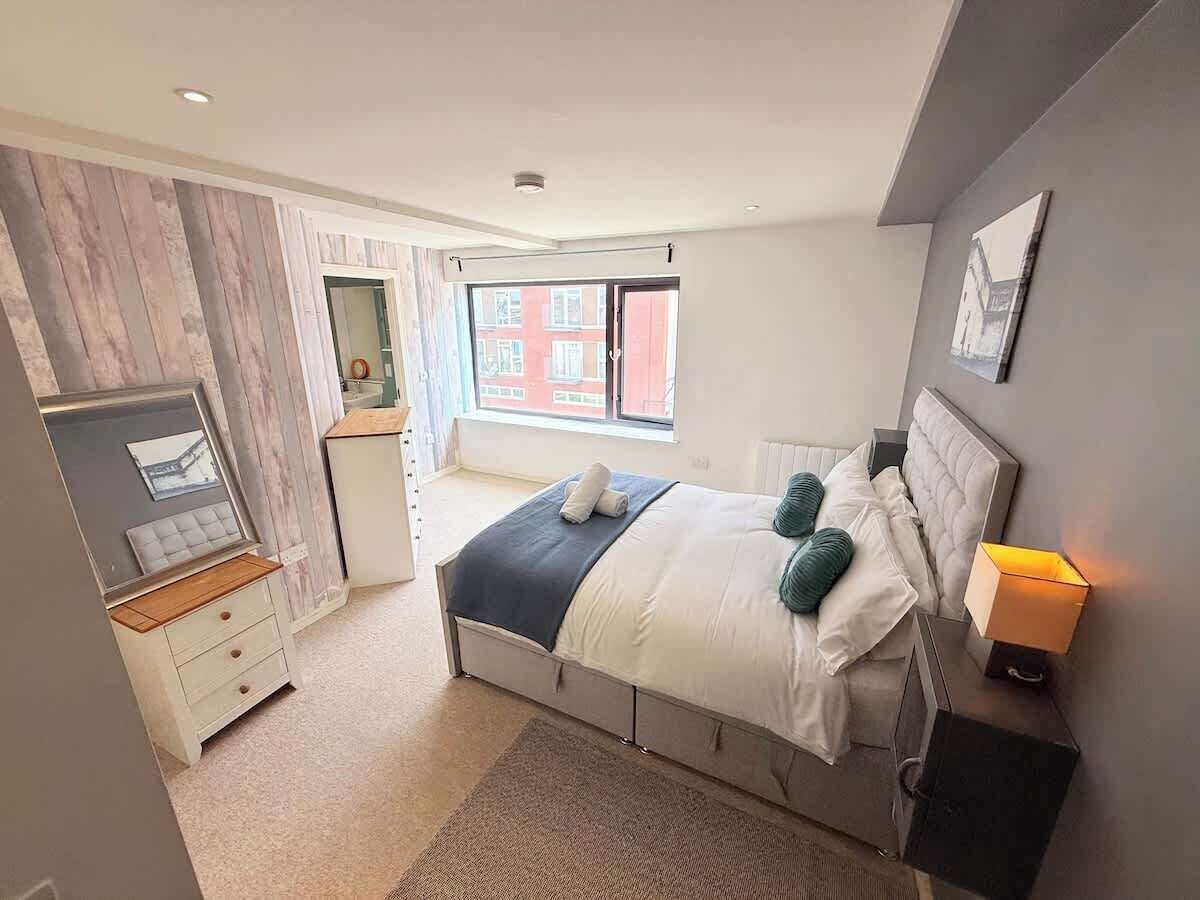 Een moderne slaapkamer met een bed, nachtkastje en raam met uitzicht op gebouwen.