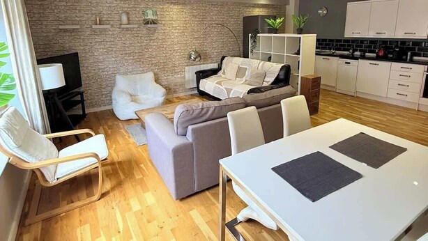 En moderne opholdsstue med spiseplads, en sofa, et fjernsyn og et køkken med lyse skabe.