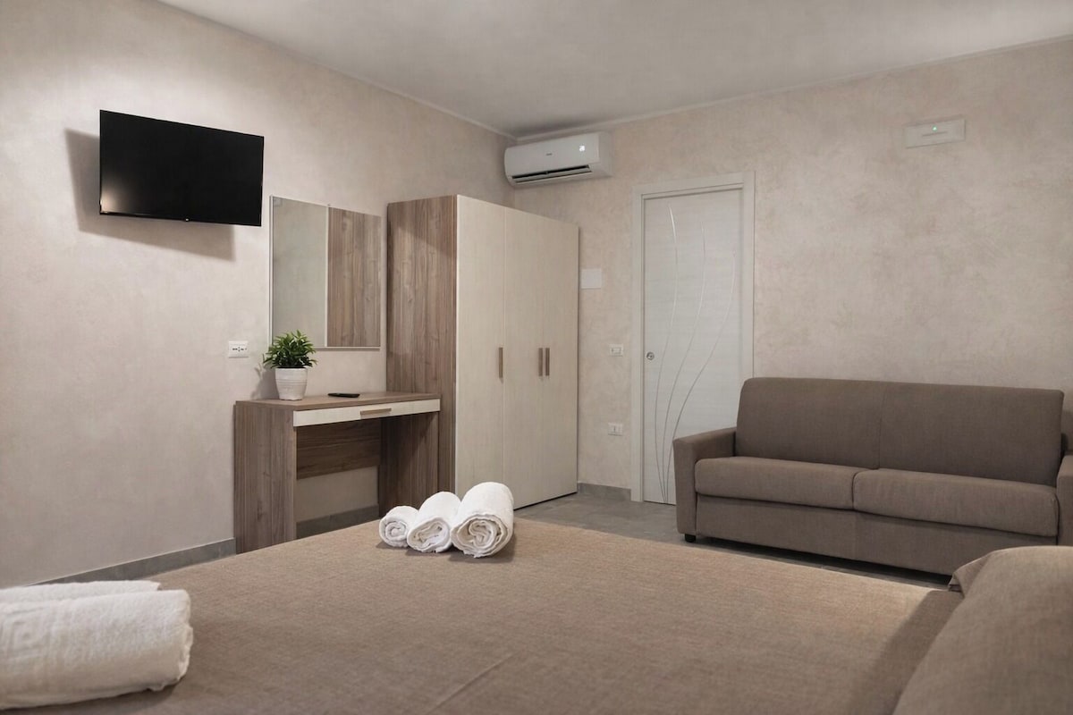 Une chambre d’hôtel moderne équipée d’un lit, d’un canapé, d’un bureau avec un miroir et d’un téléviseur à écran plat fixé au mur.