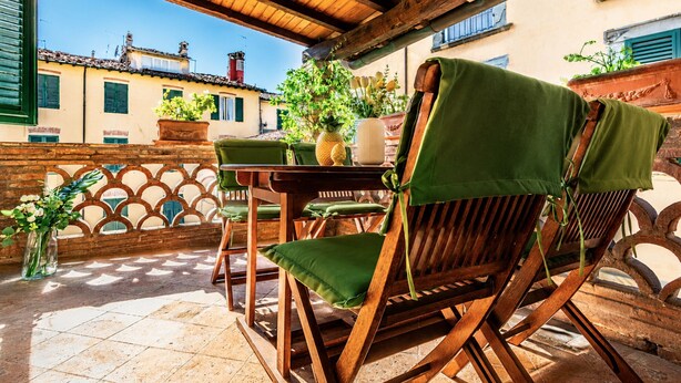 Un área de estar al aire libre con madera, que incluye una mesa, sillas, plantas en macetas y vistas de edificios al fondo.