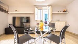 Una sala de estar moderna con una mesa de comedor de vidrio, sillas negras, un televisor de pantalla plana y una ventana con persianas azules.