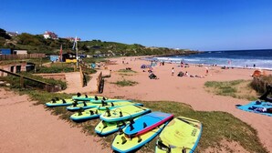 Une plage où l’on aperçoit des planches de surf alignées, un rivage sablonneux et un ciel bleu dégagé.