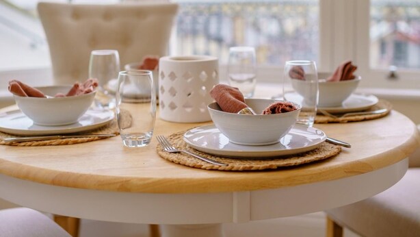 A round dining table with white plates, bowls, and cutlery, set with woven placemats and a white candle holder. - 2 Bed in Bideford (oc-77589) (Northam)
