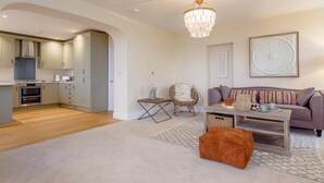 A modern living room with a sofa, ottoman, and coffee table, a dining area with a chandelier, and a kitchen with cabinets and appliances. - 2 Bed in Bideford (oc-77589) (Northam)