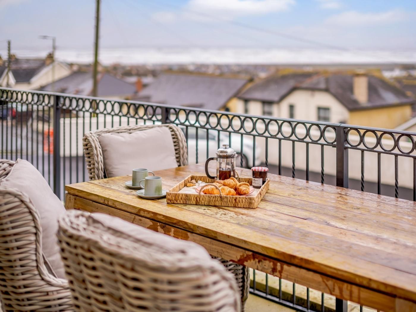 Una mesa de madera con una cafetera, croissants y un tarro de mermelada en un balcón con sillas de mimbre.