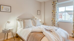 Una cama bien hecha con ropa blanca, una lámpara de noche y una mesita con lámpara. Hay dos cuadros en la pared.