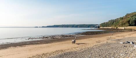 A pebbly beach with a clear sky, calm sea, and distant hills.