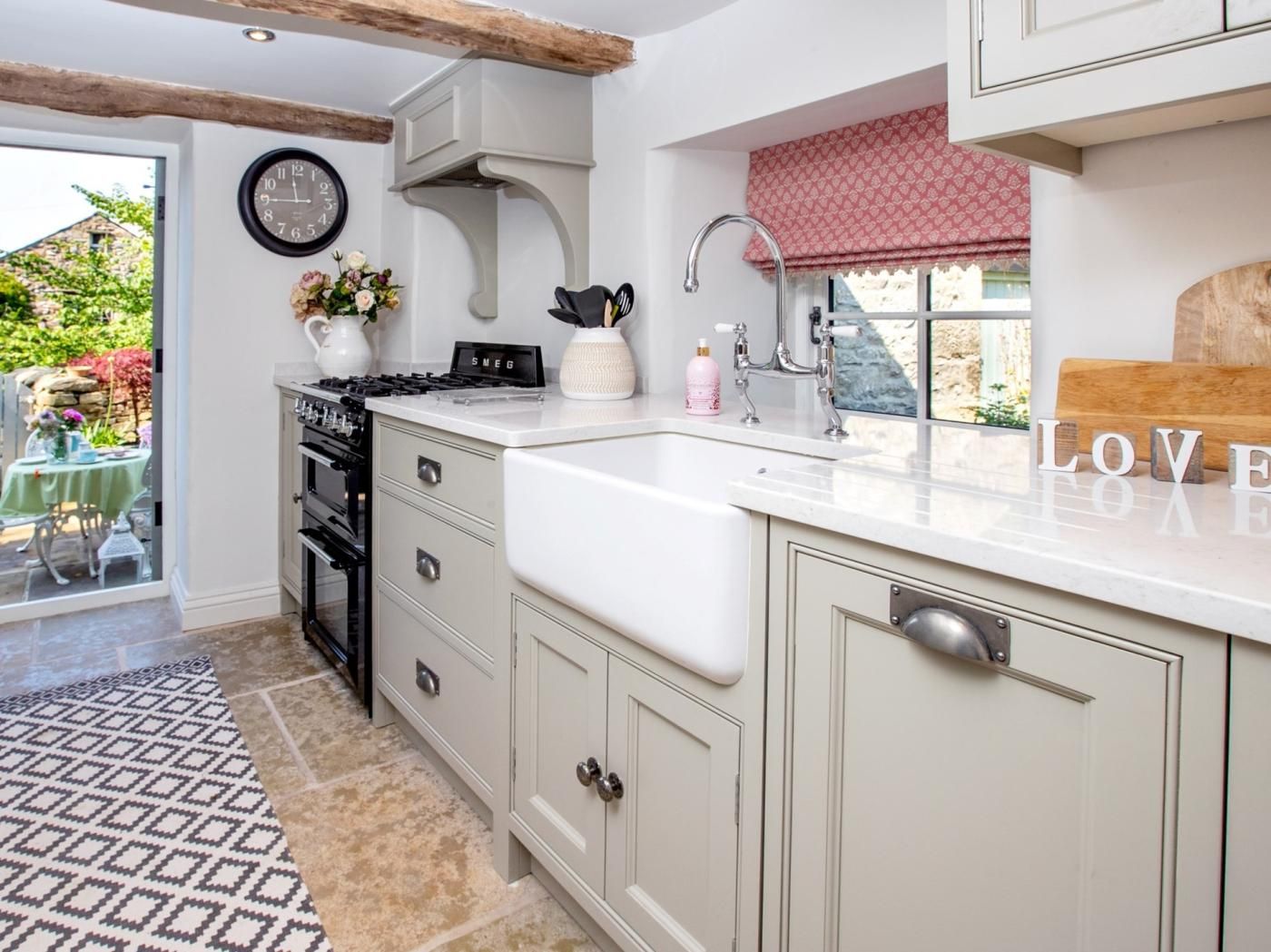 Een keuken met een ingebouwde kookplaat, een spoelbak en een raam met een rode vouwsluier. Er staat een klok aan de muur en een vaas met bloemen op het aanrecht.