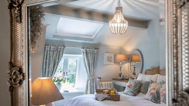 Quarto com um espelho grande e decorativo, um candelabro, cama com almofadas, mesa de cabeceira com abajur e janela com cortinas.