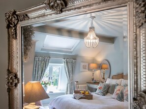 Quarto com um espelho grande e decorativo, um candelabro, cama com almofadas, mesa de cabeceira com abajur e janela com cortinas.