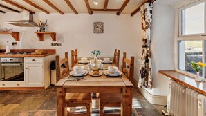 Een eetgedeelte met een houten tafel en stoelen, een keuken met een fornuis en kastjes, een klok aan de muur, en een raam met gordijnen.