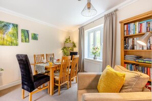 A cozy living room with a sofa, a dining table with chairs, a bookshelf, and a window with curtains. - 3 Bed in Bampton (oc-77931) (Bampton)
