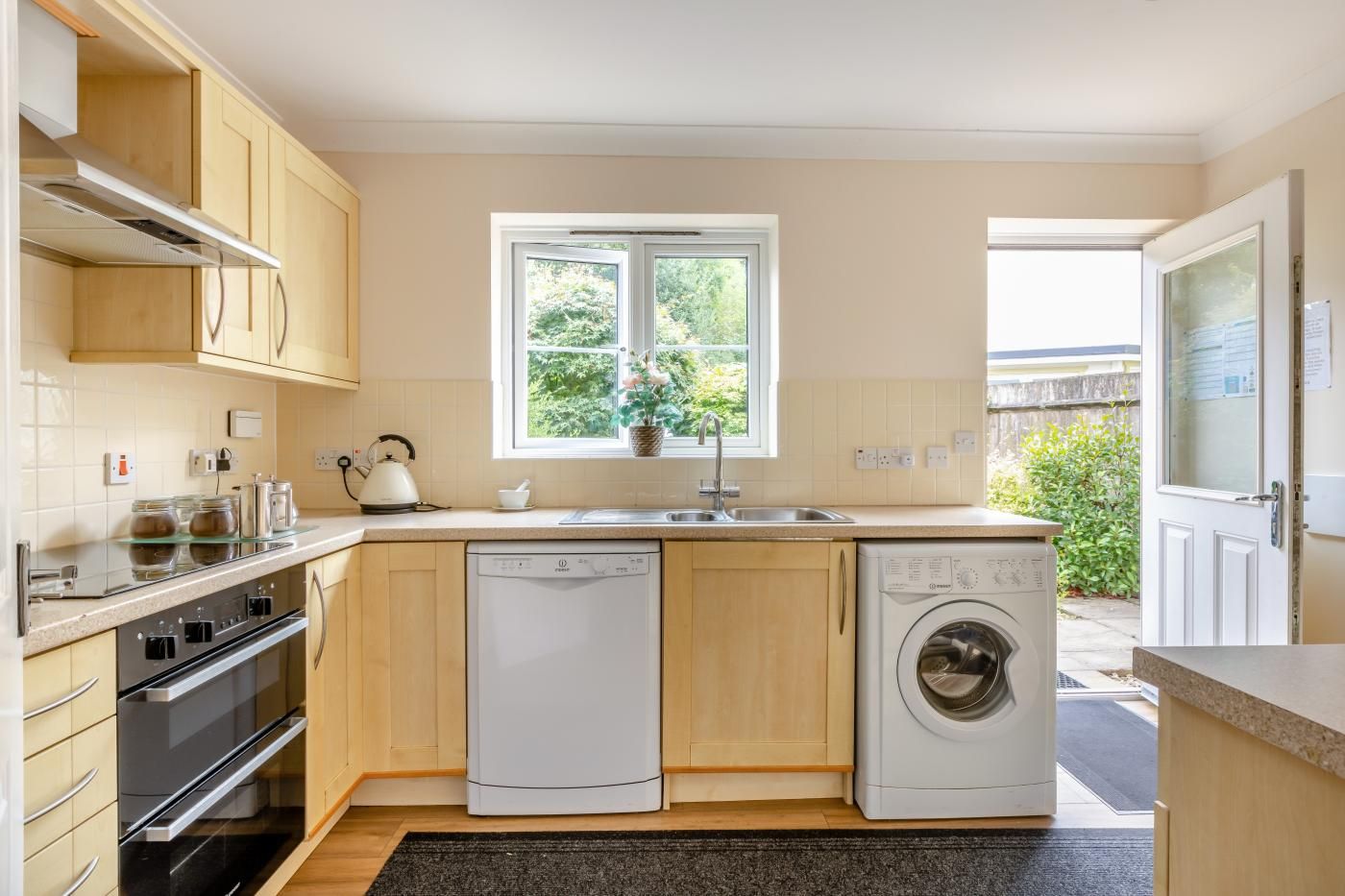 Une cuisine compacte équipée d’un lave-vaisselle, d’un four et d’un lave-linge, avec des meubles en bois et une fenêtre donnant sur un espace verdoyant.