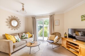 A living room with a sofa, coffee table, wicker chair, TV stand, and a large window with curtains. - 3 Bed in Bampton (oc-77931) (Bampton)