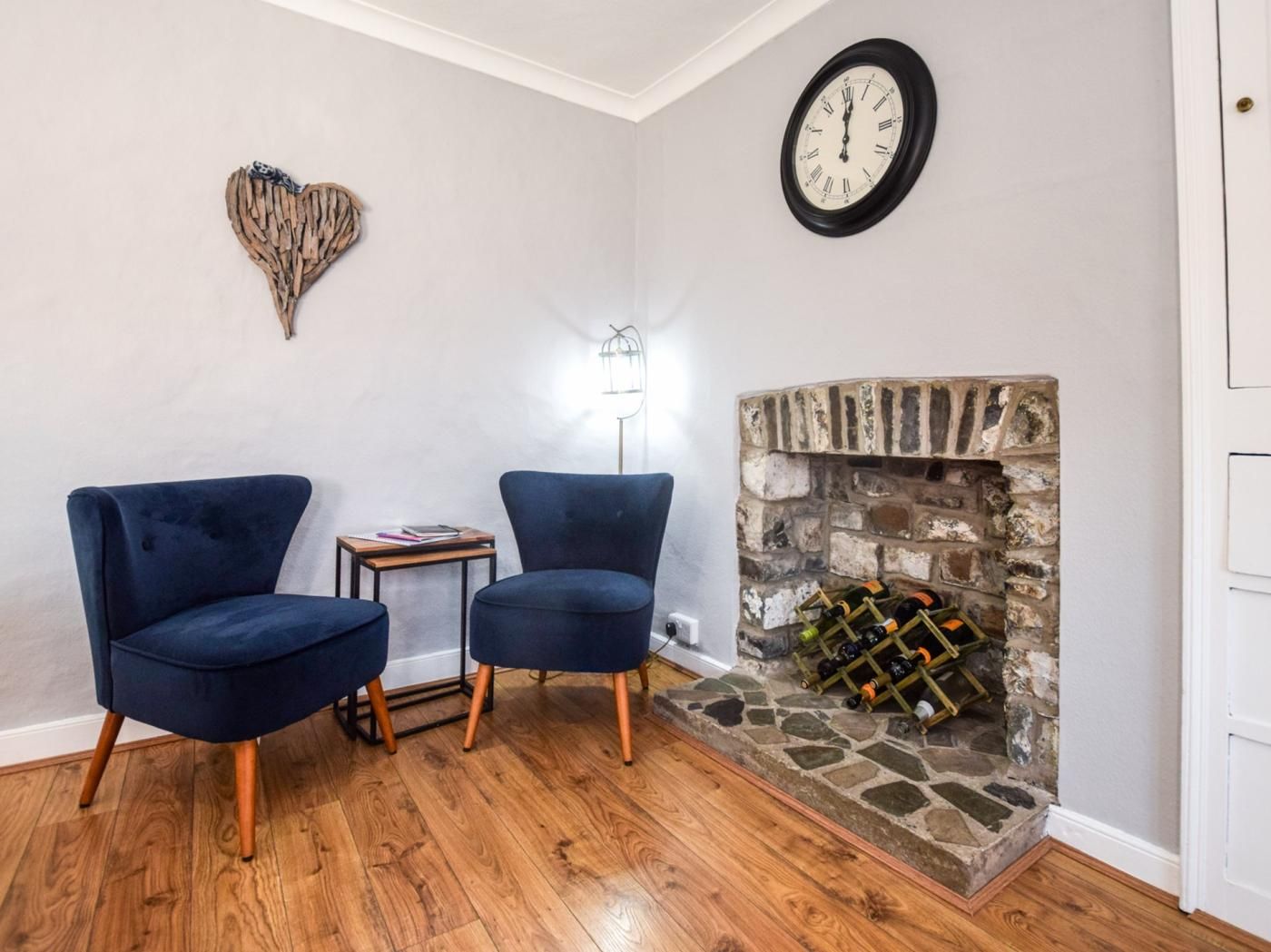 A room with two blue armchairs, a small table, a wall clock, a decorative heart, a floor lamp, and a stone fireplace with wine bottles.