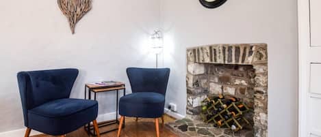 A room with two blue armchairs, a small table, a wall clock, a decorative heart, a floor lamp, and a stone fireplace with wine bottles.