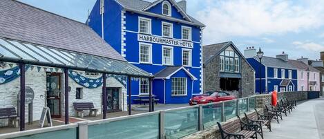 A blue building labeled 'Harbourmaster Hotel' with a stone facade, a covered seating area, and a row of benches along a waterfront.