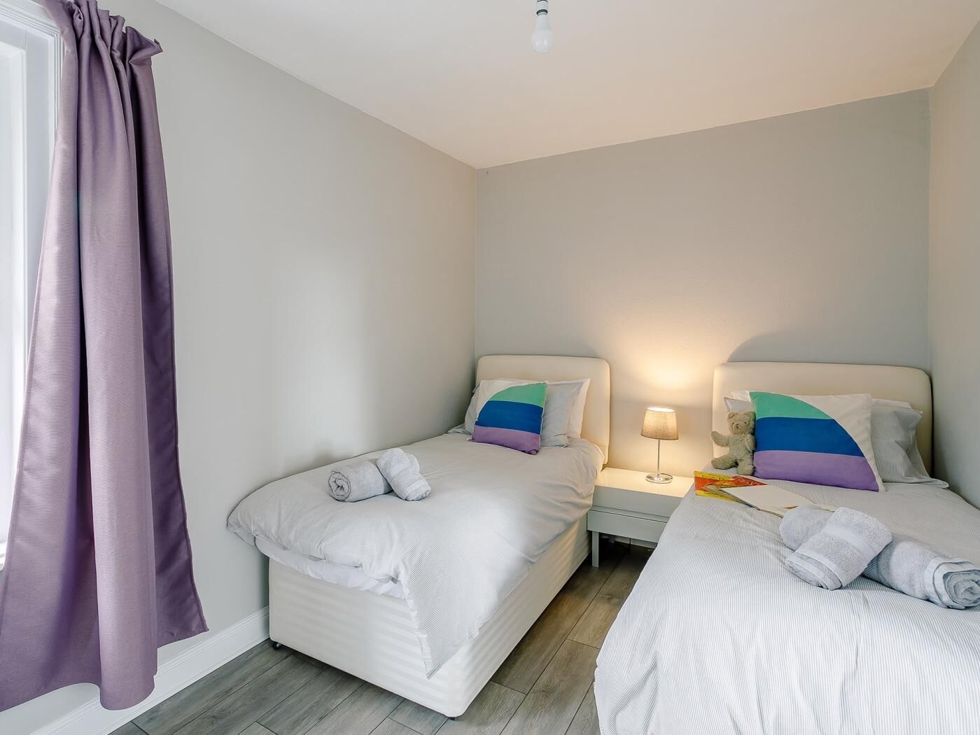 A hotel room with two beds, a nightstand, a lamp, and a teddy bear.