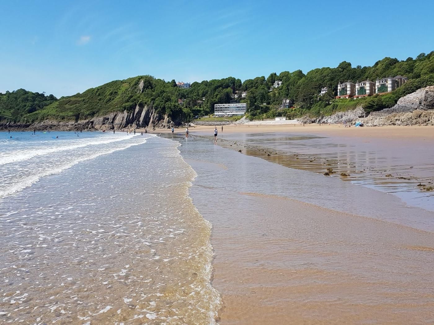 A beach with clear water, a sandy shore, and buildings in the background.
